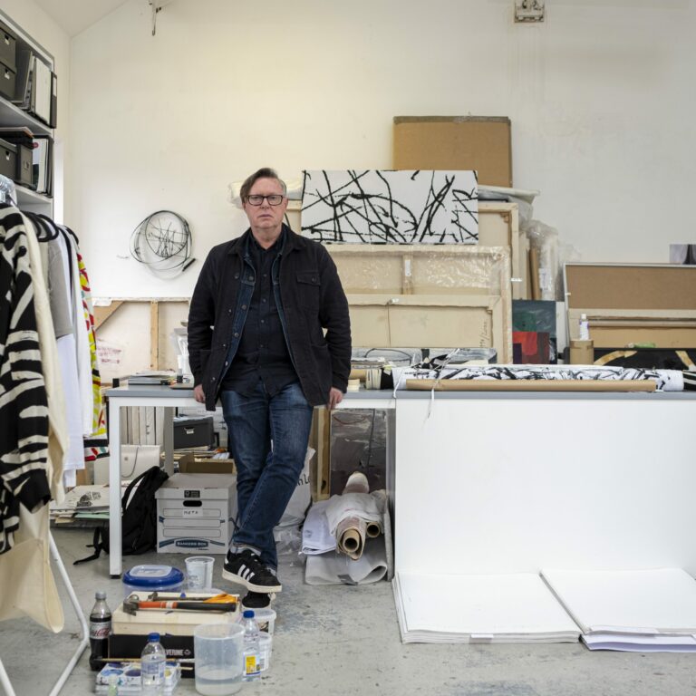A man who lives in Partick standing in the middle of a studio, with artwork on the left.